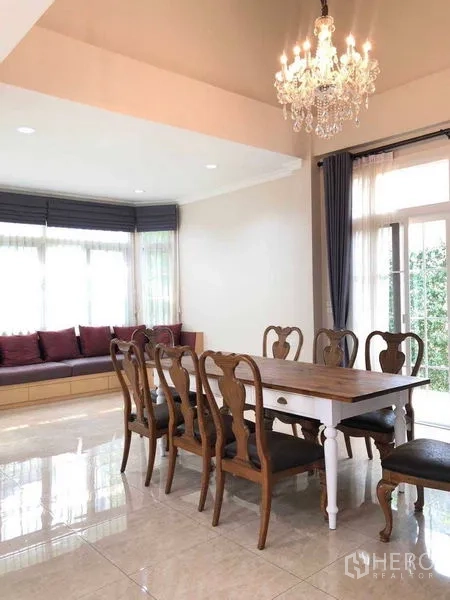 บ้าน ให้เช่าในBang Na, Bangkok - ห้องทานอาหารสว่าง โต๊ะไม้ยาว โคมระย้า และม้านั่งริมหน้าต่างมองสวน