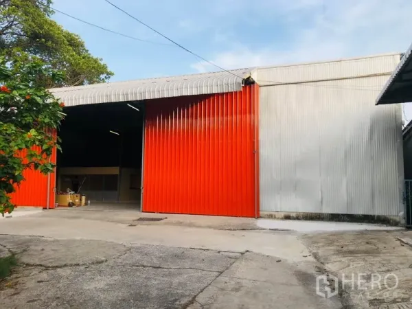 Warehouse for rent in Wang Thonglang, Bangkok - Front façade of the warehouse with red metal doors, driveway access and partial tree shade.