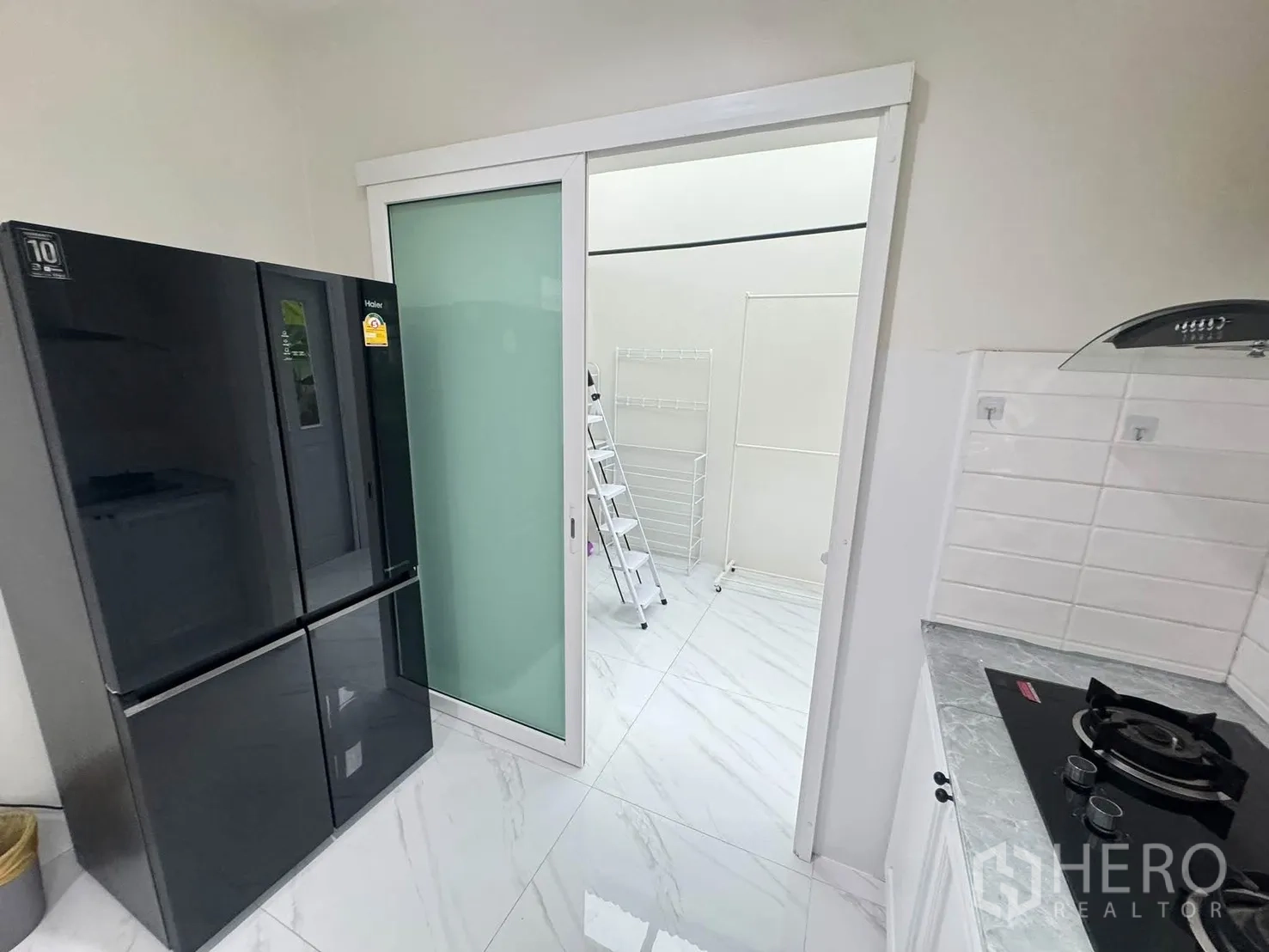 Detached House for rent or sale in Bang Phli, Samut Prakan - Kitchen entry with large black refrigerator and sliding frosted glass door to a utility area with drying rack.