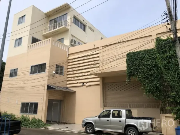 工厂 出租或出售在Bang Lamung, Chon Buri - 米色工业建筑外立面，带装卸区、卷闸门和停靠的皮卡车。