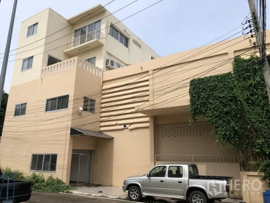 Factory for rent or sale in Bang Lamung, Chon Buri - Front exterior of a beige industrial building with loading bay, roller shutter and a parked pickup truck.