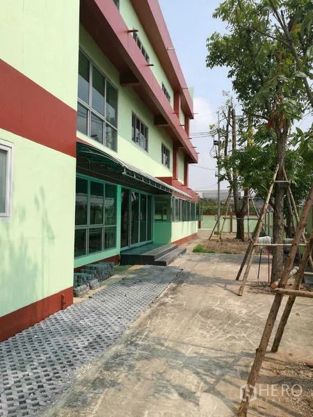 Factory for rent in Mueang Samut Sakhon, Samut Sakhon - Side facade of the factory with shaded walkway, windows, and young trees.