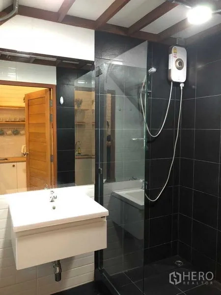 Detached House for rent in Phra Nakhon, Bangkok - Modern bathroom with black tiles, glass shower enclosure, and water heater next to a white vanity.