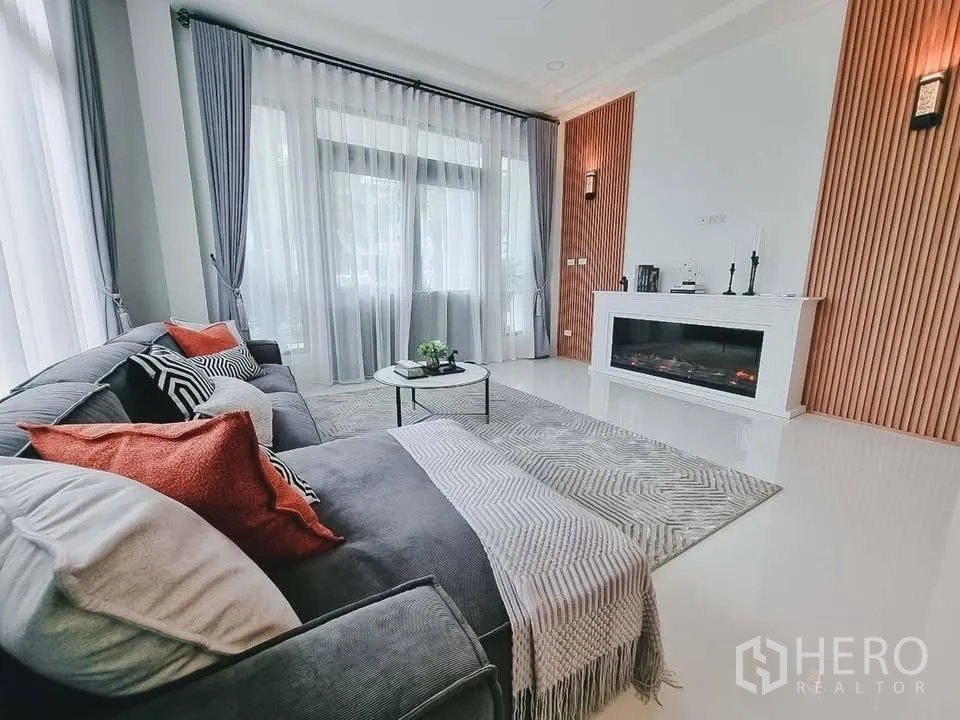 独栋房屋 for rent or sale in Bang Phli, Samut Prakan - 明亮客厅，落地窗与纱帘，灰色转角沙发和现代装饰壁炉。