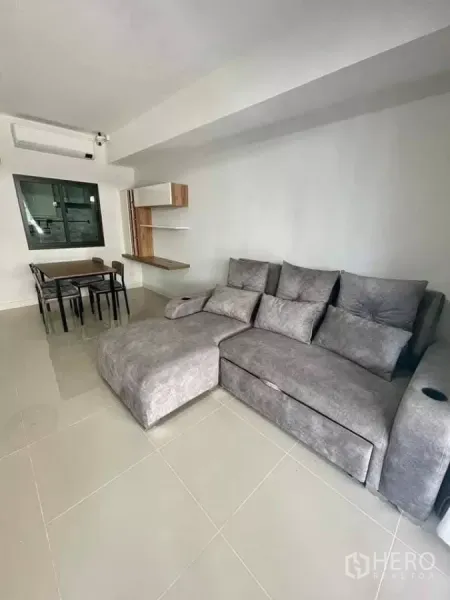 Townhouse for rent in Bang Phli, Samut Prakan - Bright living room with large gray sectional sofa, tiled floor, dining table and floating shelves.