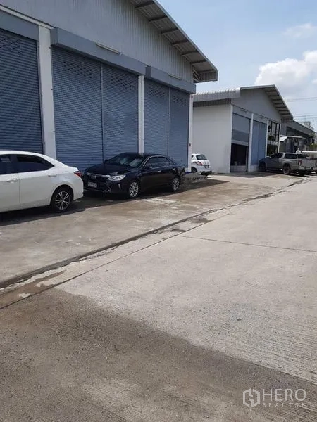 Warehouse for rent in Bang Phli, Samut Prakan - Wide driveway in front of warehouse units with loading access and parked cars.