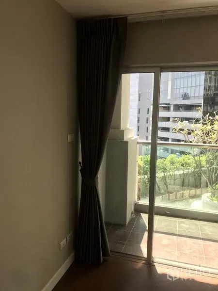 Condominium for rent or sale in Huai Khwang, Bangkok - Sliding glass door to a balcony with city view and greenery beside dark blackout curtains.
