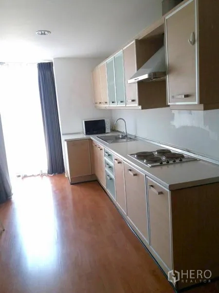 Condominium for rent in Watthana, Bangkok - Bright galley kitchen with long countertop, light wood cabinets, stovetop and range hood beside a windowed door.