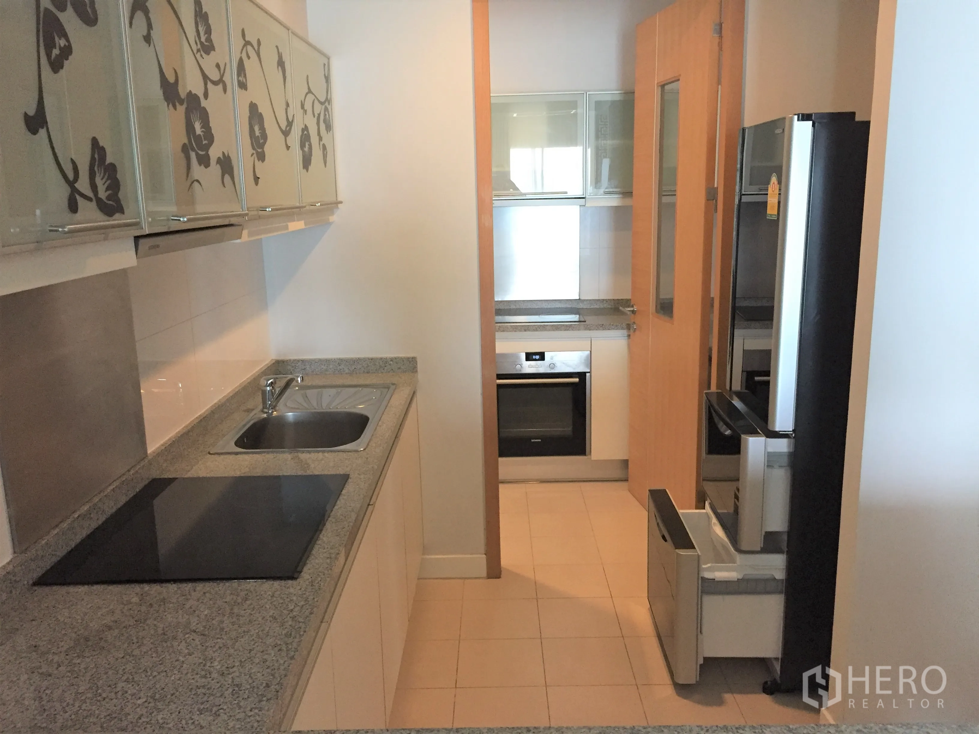 Condominium for rent or sale in Khlong Toei, Bangkok - Galley kitchen with granite countertops, electric cooktop, built-in oven and floral glass cabinets.