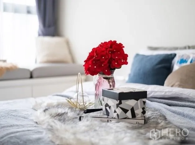 Condominium for sale in Khlong Toei, Bangkok - Bedroom decor close-up with red flowers and accessories on a grey duvet.