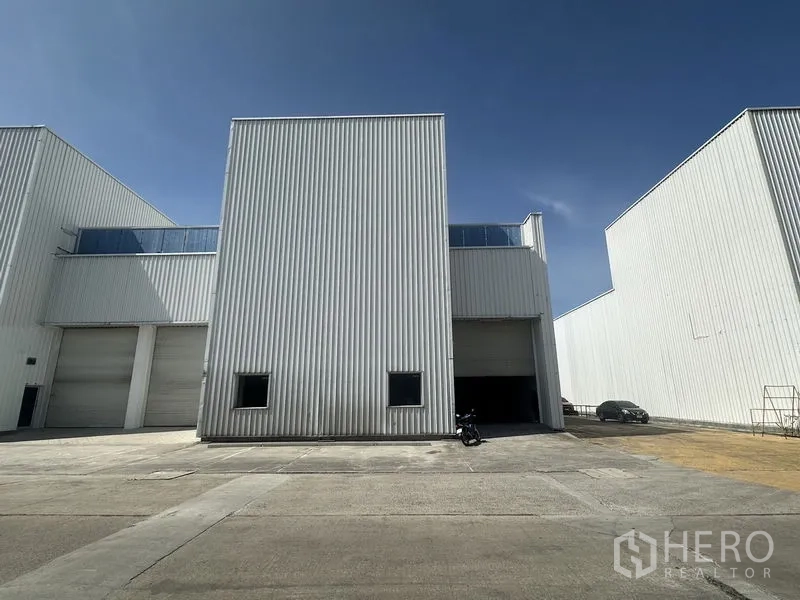 Factory for rent in Khlong Luang, Pathum Thani - Front view of a white corrugated factory unit with a large roller shutter and wide driveway under a blue sky.