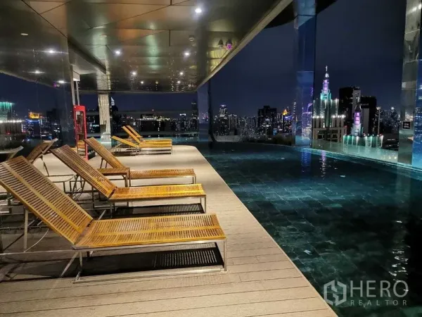 Condominium for rent in Ratchathewi, Bangkok - Rooftop infinity pool at night with sun loungers and the Bangkok skyline.