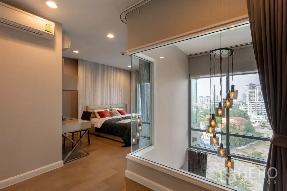 Condominium for rent or sale in Khlong Toei, Bangkok - Bedroom with city-view windows, an interior glass wall and hanging pendant lights.