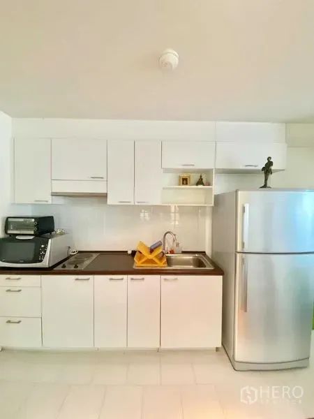 Condominium for rent in Watthana, Bangkok - Bright compact white kitchen with upper and lower cabinets, cooktop, sink, and a stainless refrigerator.