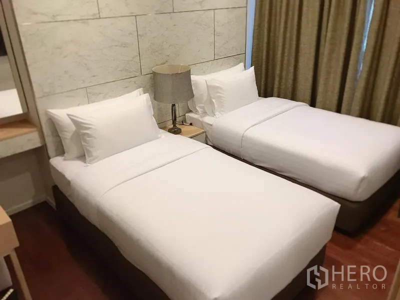 Condominium for rent in Watthana, Bangkok - Second bedroom with two single beds, bedside lamp and a paneled feature wall with curtains.