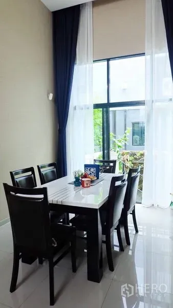 Townhouse for rent or sale in Suan Luang, Bangkok - Dining area with double-height ceiling and tall glass windows overlooking greenery.
