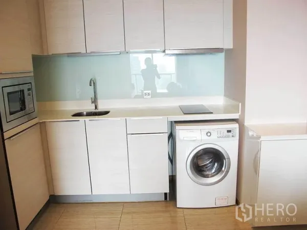 Condominium for rent in Watthana, Bangkok - Compact light-wood kitchen with built-in microwave, sink, glass backsplash, induction hob and front-load washing machine.