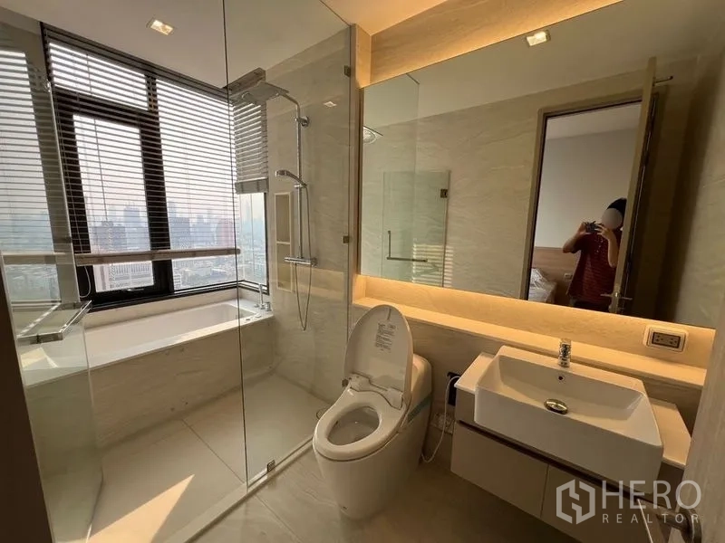 Condominium for rent in Ratchathewi, Bangkok - Bathroom with bathtub by the window, glass shower, vanity and toilet overlooking the city.