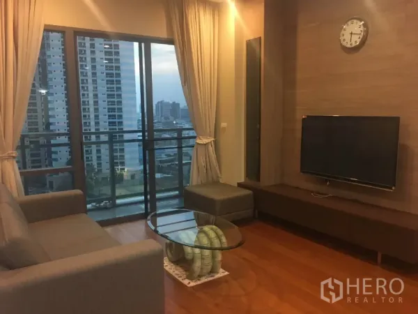 Condominium for rent in Khlong Toei, Bangkok - Warm living room with sofa, TV wall and floor-to-ceiling windows opening to a city-view balcony.