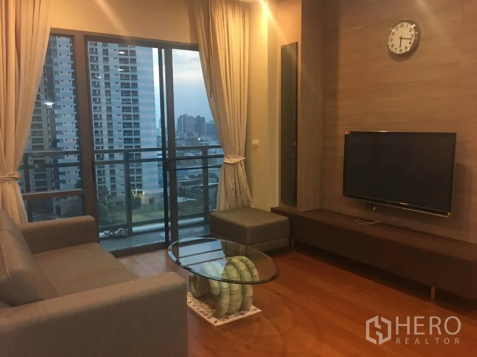 Condominium for rent in Khlong Toei, Bangkok - Warm living room with sofa, TV wall and floor-to-ceiling windows opening to a city-view balcony.