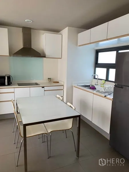 Condominium for rent in Watthana, Bangkok - Kitchen with window, sink, upper cabinets, refrigerator and a white dining table with metal legs.