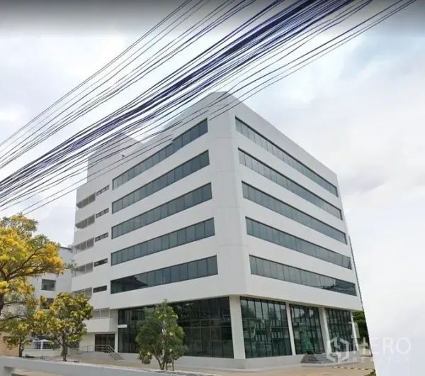 Office Space for rent in Din Daeng, Bangkok - Corner view of a modern white mid-rise office building with long horizontal windows, glassy ground floor, trees, and a paved driveway.