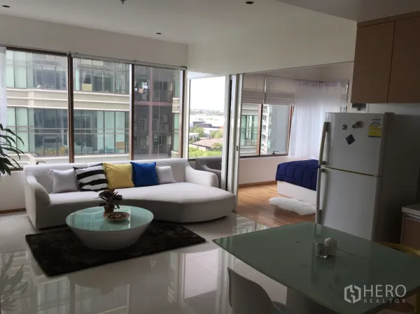 Condominium for rent in Khlong Toei, Bangkok - Bright open‑plan living area with white sofa, glass coffee table, dining corner and large windows with city views.