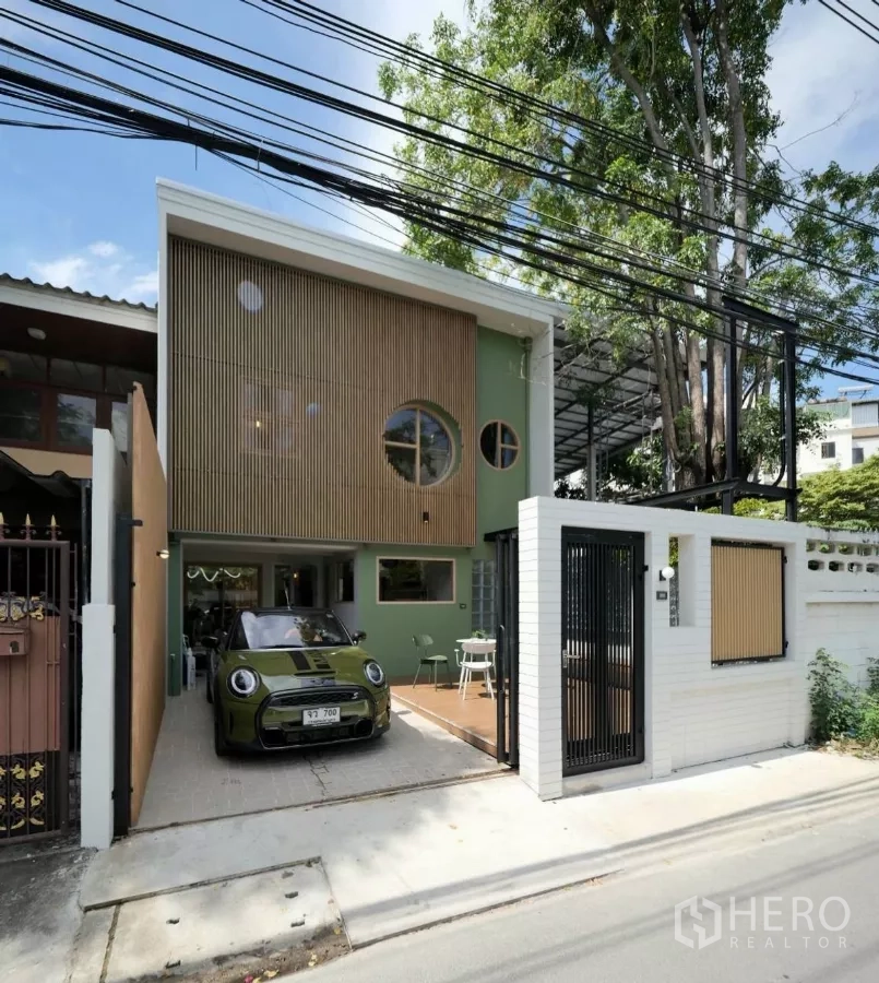 Townhouse for rent in Watthana, Bangkok - Daytime exterior showing driveway parking, round windows, slatted wood screen and a white brick wall with sliding gate.