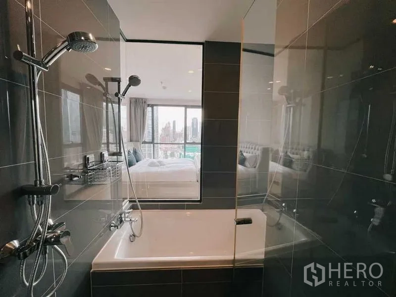 Condominium for rent in Ratchathewi, Bangkok - Modern bathroom with bathtub and handheld shower, window to bedroom and city view.