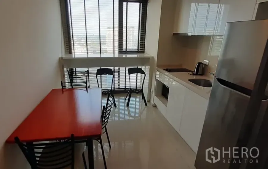 Condominium for rent in Khlong Toei, Bangkok - Compact kitchen with glossy white cabinetry, stainless refrigerator, and a dining table by large windows.