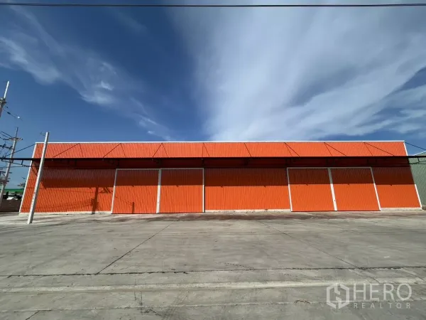 Warehouse for rent in Khlong Toei, Bangkok - Wide orange warehouse facade with multiple large loading doors and a broad concrete yard under a blue sky.