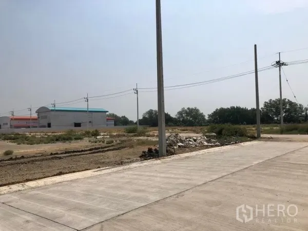 Land for sale in Mueang Samut Sakhon, Samut Sakhon - Corner of a concrete road with utility poles, rubble pile, and factory buildings in the background.