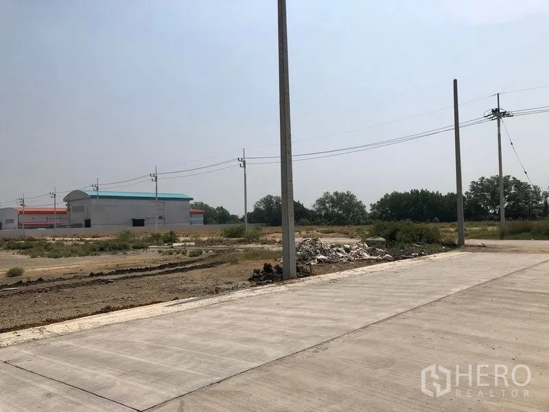 Land for sale in Mueang Samut Sakhon, Samut Sakhon - Corner of a concrete road with utility poles, rubble pile, and factory buildings in the background.