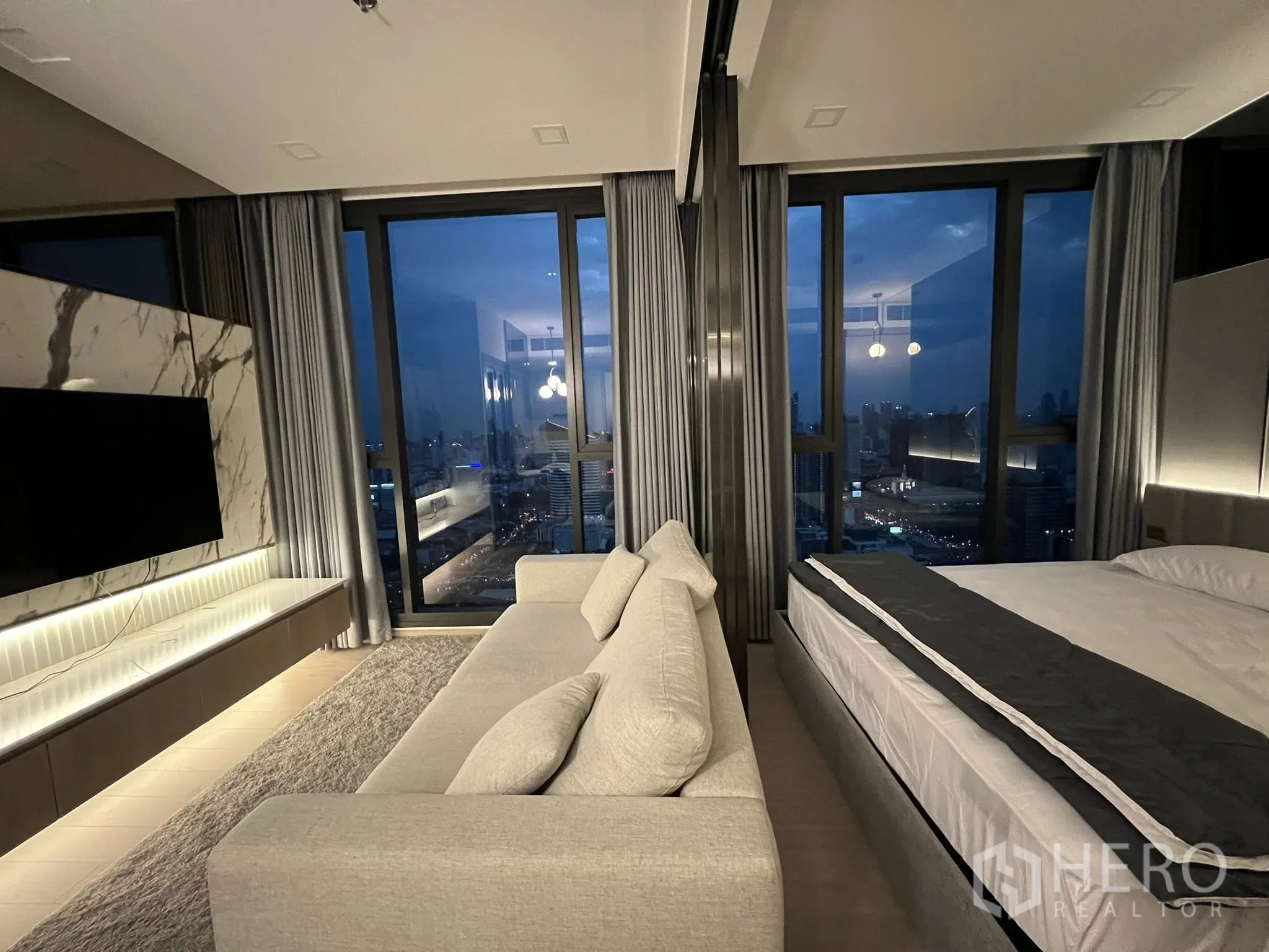 คอนโด for rent in Huai Khwang, Bangkok - ห้องรับแขกและห้องนอนกั้นกระจก ชั้นสูง กระจกบานใหญ่ ผนังทีวีลายหินอ่อน โซฟา และวิวเมืองยามค่ำคืน