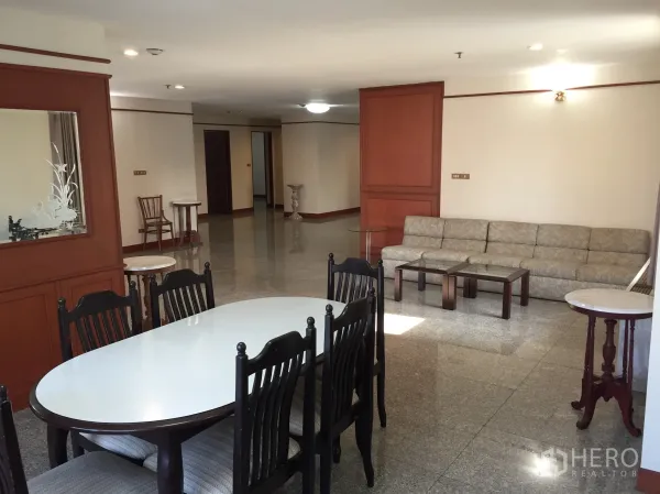 Condominium for rent in Watthana, Bangkok - Dining zone with oval table and six chairs beside a long sofa, wood paneling and glossy floors in a large condo living room.