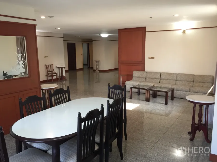 Condominium for rent in Watthana, Bangkok - Dining zone with oval table and six chairs beside a long sofa, wood paneling and glossy floors in a large condo living room.
