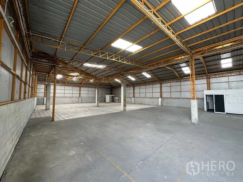 Warehouse for rent in Mueang Pathum Thani, Pathum Thani - Open warehouse interior with high steel truss roof, skylights, and a concrete floor with a tiled section.