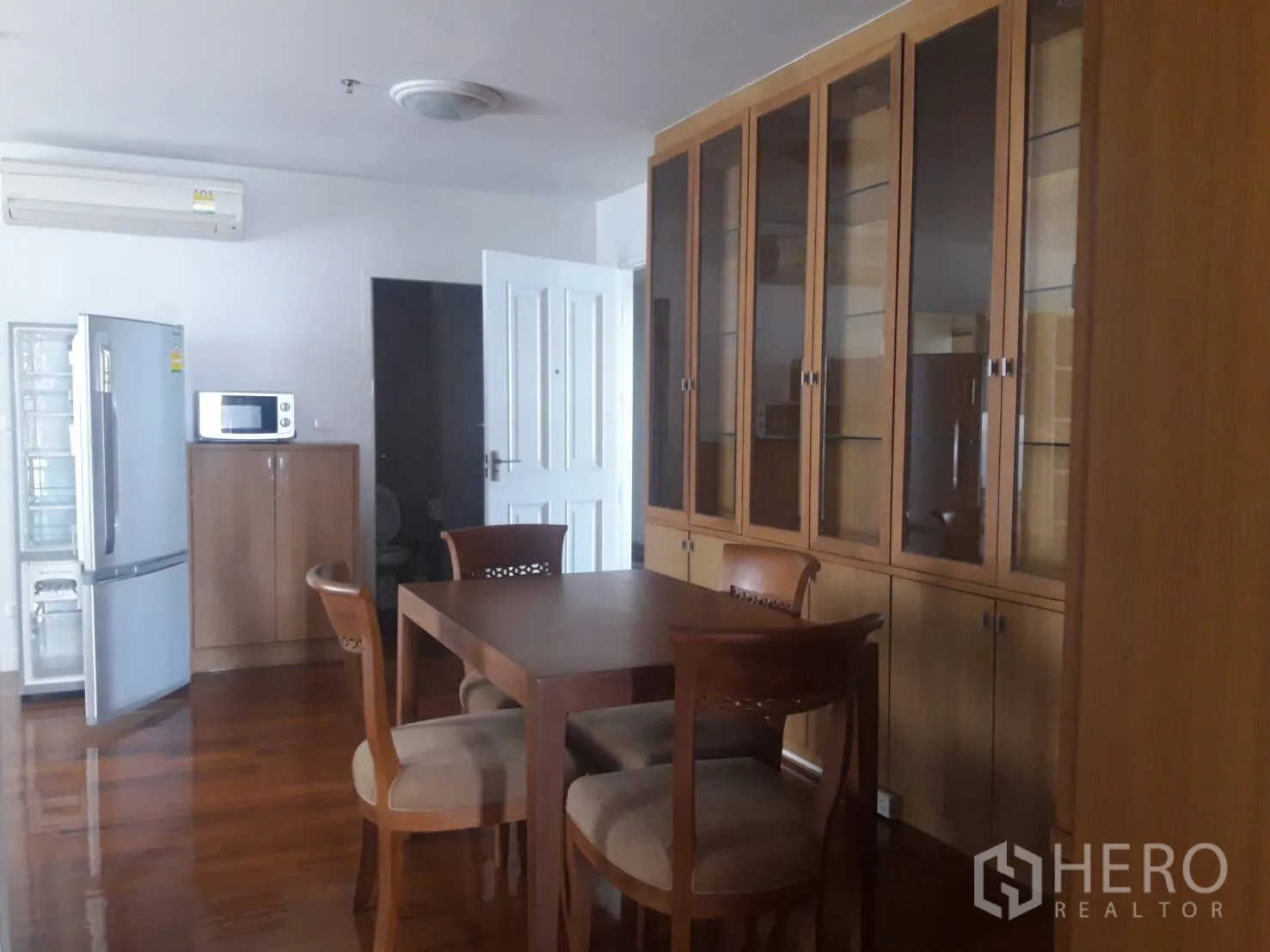 Condominium for rent in Khlong Toei, Bangkok - Dining area with wooden table and chairs, tall glass-front cabinets, refrigerator and microwave on glossy wood floors.
