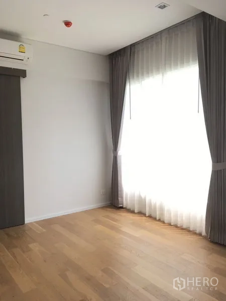 Condominium for rent or sale in Chatuchak, Bangkok - Sunlit empty room with a large curtained window, air conditioner and wood floors.