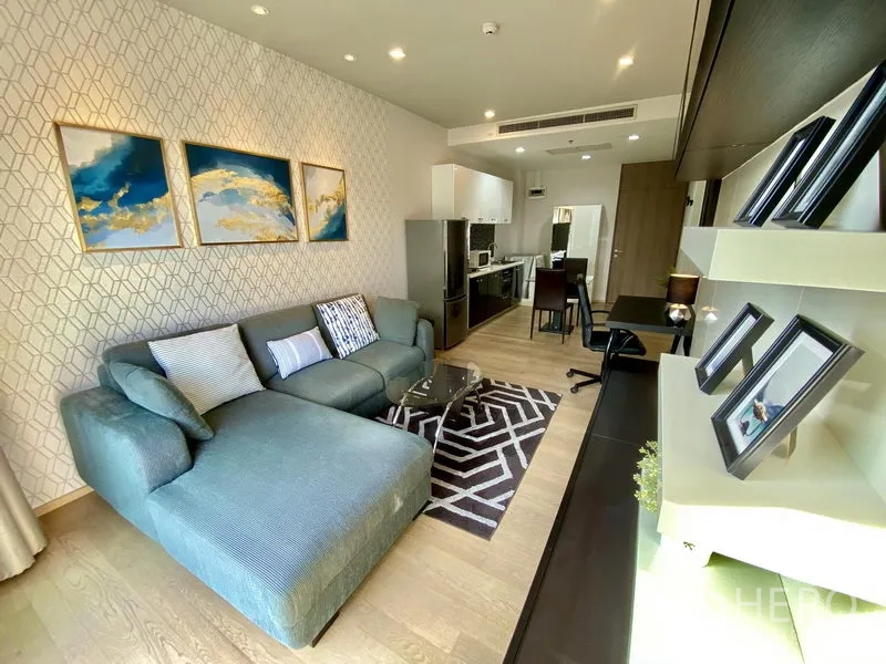 Condominium for rent in Bangkok - Open-plan living room with teal sectional sofa, wall art, built-in shelving and a kitchenette.