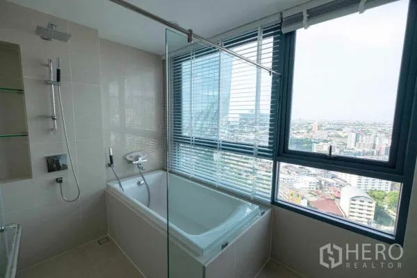 公寓 出租在Bang Na, Bangkok - 现代卫浴，浴缸靠大窗，玻璃淋浴隔断与顶喷花洒，可观城市天际线。