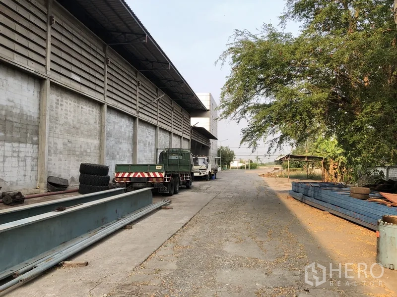 Factory for rent in Bang Bua Thong, Nonthaburi - Side yard beside the factory with trucks, steel beams, and mature trees.