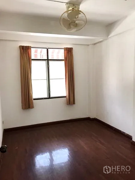 Townhouse for rent in Khlong Toei, Bangkok - Simple bedroom with window, wall fan and glossy wood floors.