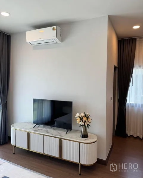 Detached House for rent or sale in Bang Phli, Samut Prakan - TV on a marble-top console below a wall-mounted air conditioner beside window curtains.