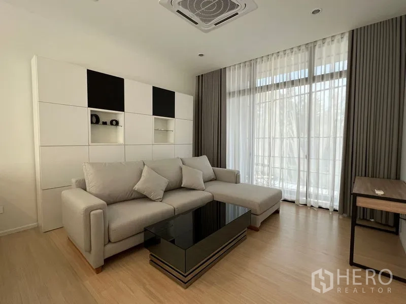 Townhouse for rent in Suan Luang, Bangkok - Light-filled living room with an L-shaped sofa, built-in shelving, large windows and a black coffee table.