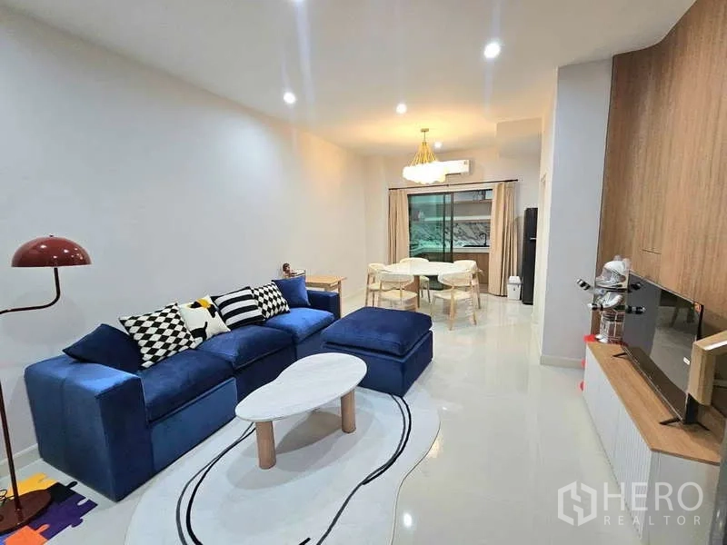 Townhouse for rent in Suan Luang, Bangkok - Open-plan living and dining area with blue sectional sofa, TV wall and sliding glass doors.
