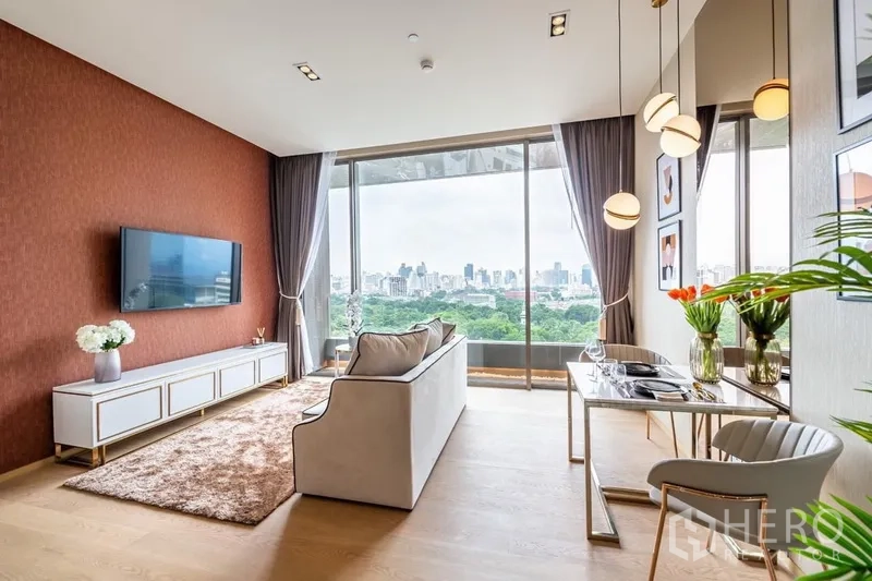 Condominium for rent or sale in Bang Rak, Bangkok - Stylish living room with sofa, wall-mounted TV, dining nook and pendant lights overlooking greenery and the Bangkok skyline.