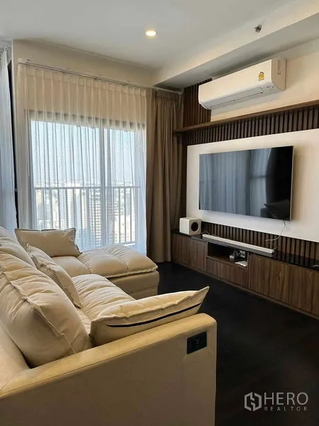 Condominium for rent in Khlong Toei, Bangkok - Sunlit living area with wall-mounted TV, air conditioner and sheer curtains by balcony.