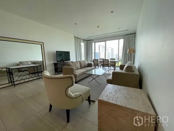 Condominium for rent in Pathum Wan, Bangkok - Bright living room with beige sofas, glass coffee table, TV console and dining set beside floor‑to‑ceiling windows with city views.