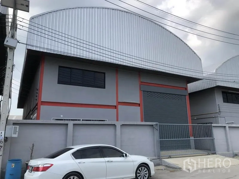 คลังสินค้า ให้เช่าในNonthaburi - ภายนอกโกดังโทนเทา-แดง หลังคาเมทัลชีททรงโค้ง ประตูม้วนขนาดใหญ่และทางเข้ามีกำแพงรั้วบนถนนคอนกรีตในย่านอุตสาหกรรม